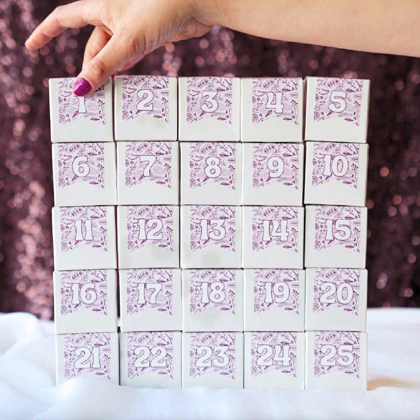 Hand with purple nail polish holding a purple and white 25-day canadian advent calendar with a foliage pattern.