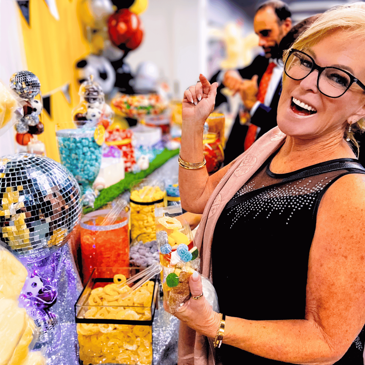 A smiling woman in formal attire holding a bag filled with colourful candy at a decorated candy buffet.