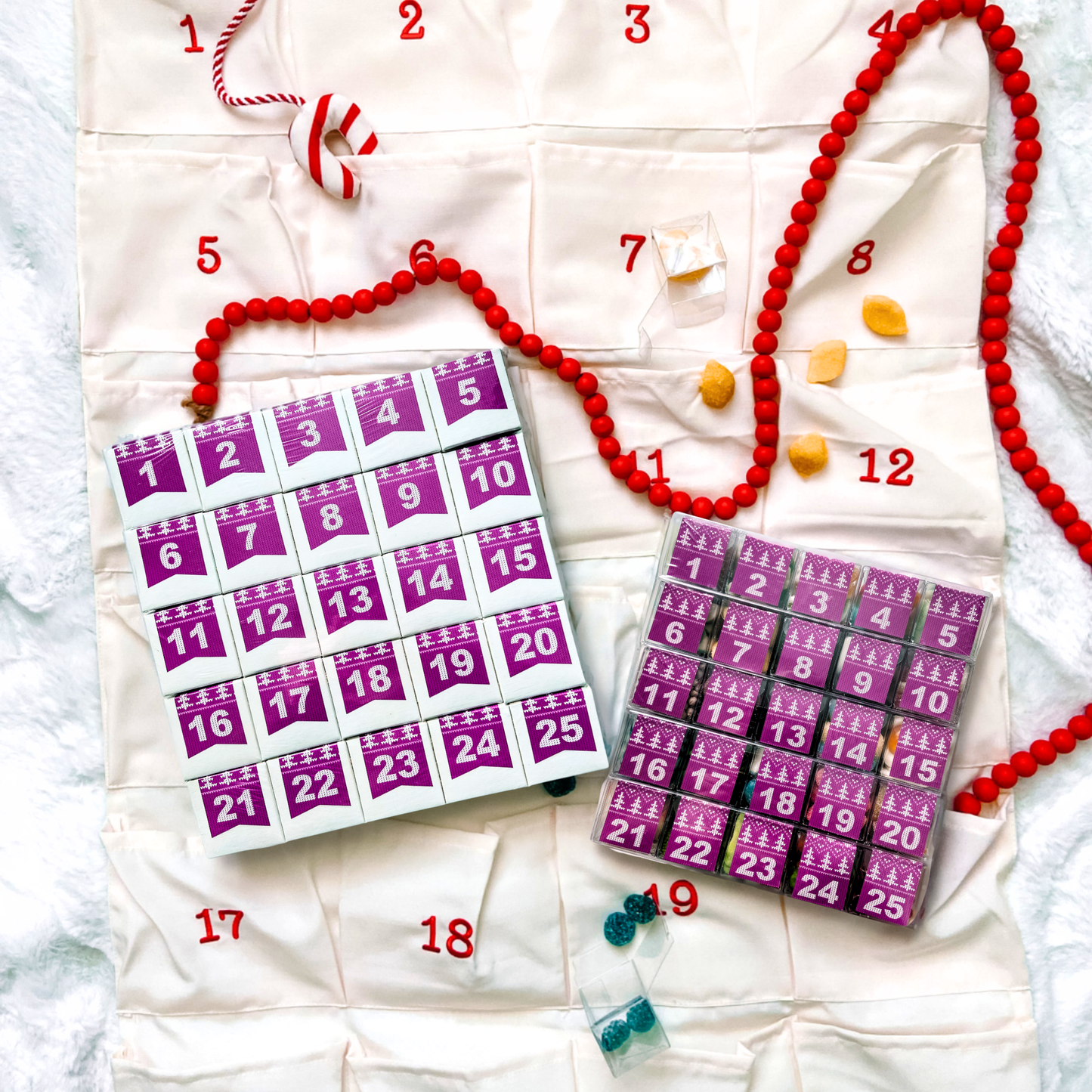 Two purple 25-day countdown calendars sitting on top of a fabric hanging pocket advent calendar decorated with a red bead garland and candy.