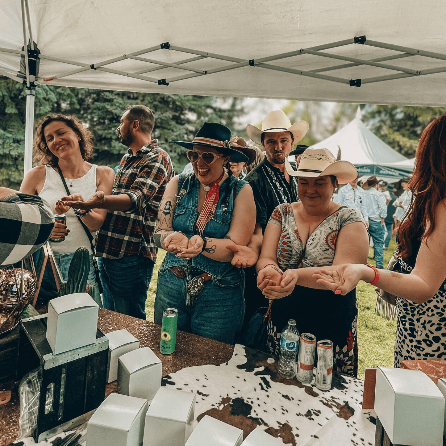 A group of people dressed in Western attire and cowboy hats stand under a white event tent with their eyes closed and hands held out, laughing during a blind candy tasting experience.