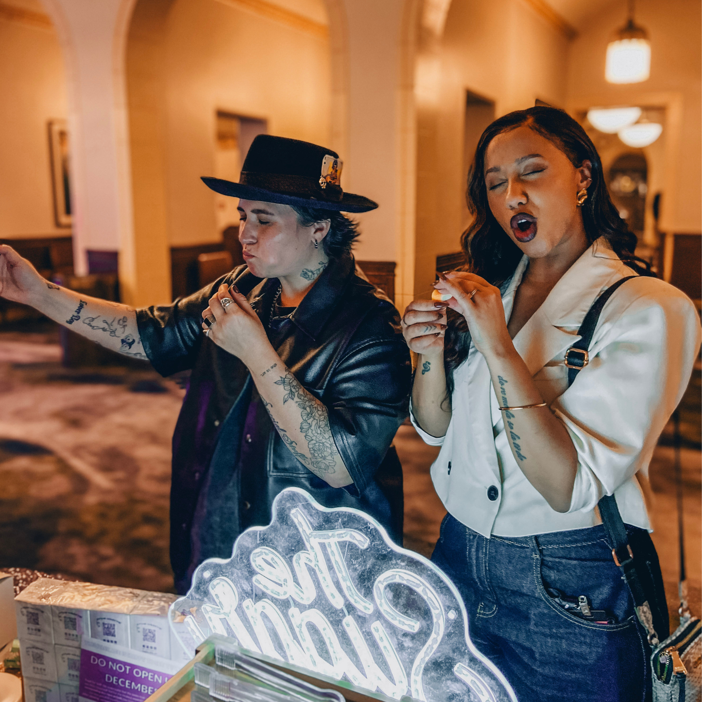 Two adults with closed eyes making playful faces while tasting candy at an indoor event.