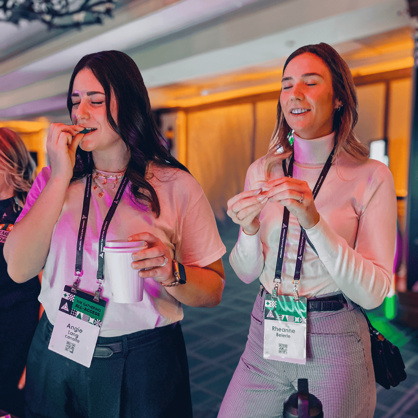 Two women wearing conference lanyards stand indoors, smiling with their eyes closed as they sample treats during a blind candy tasting event.